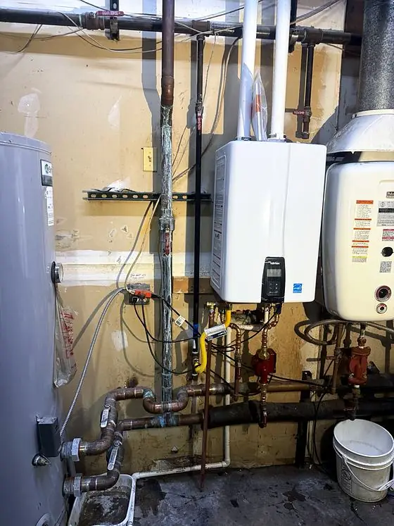 Wall-mounted tankless water heater with white vent pipes, a yellow gas connector and copper water piping in a mechanical room.