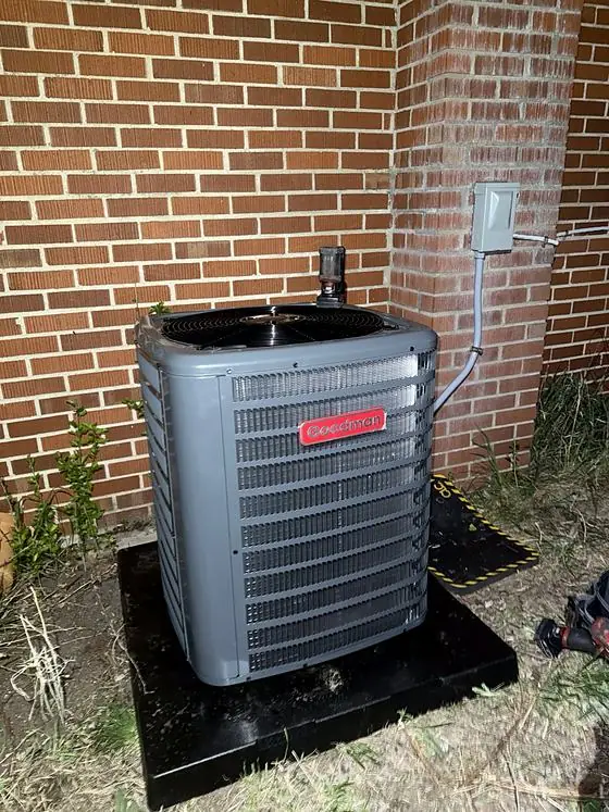 Outdoor air conditioning condenser on a black pad at the corner of a brick wall, photographed at night with a disconnect box mounted above.
