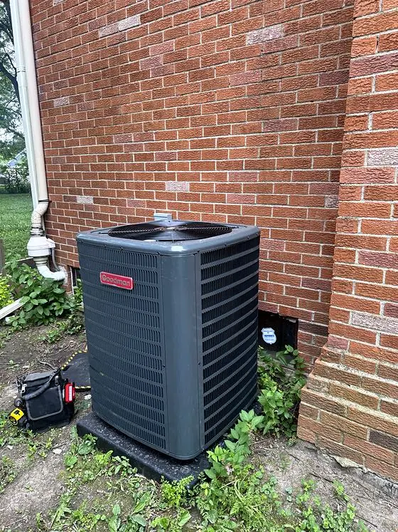 Outdoor air conditioning condenser on a level pad beside a red brick wall, with a technician's tool bag nearby.