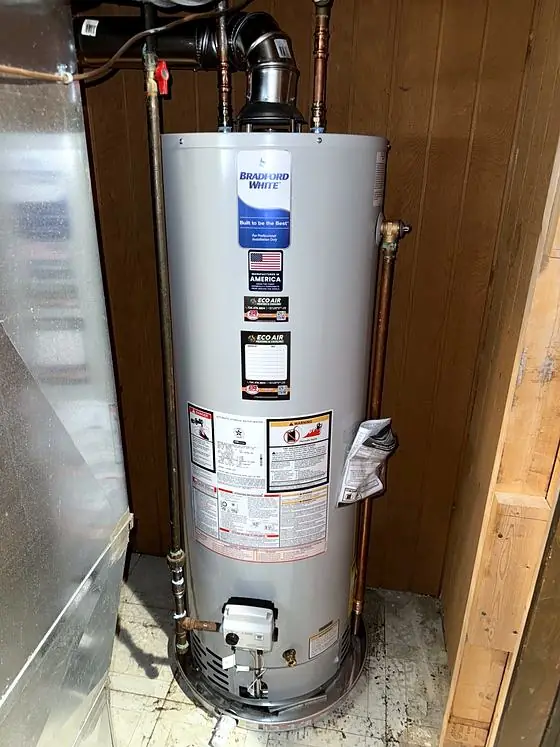 Gas tank water heater with copper supply lines and metal vent pipe, installed in a wood-paneled utility space and labeled with an Eco Air service sticker.
