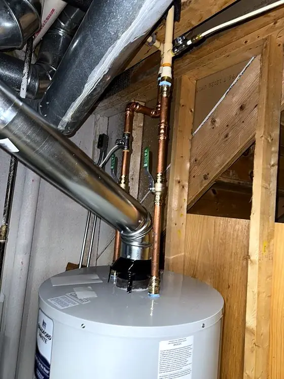 Top of a tank water heater showing copper water piping with green-handled shutoff valves and a metal vent connector under wood framing.