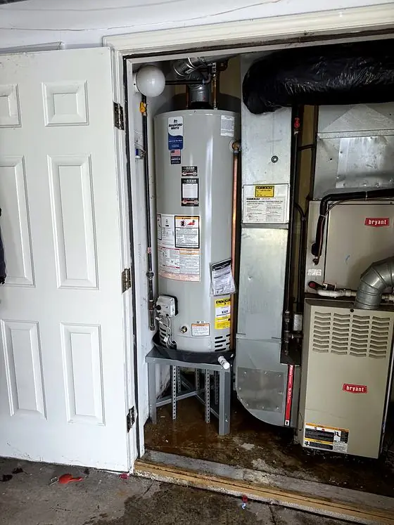 Equipment detail: gas tank water heater on a raised metal stand with a drain pan, inside a utility closet next to a furnace and ductwork. Water is visible on the floor.