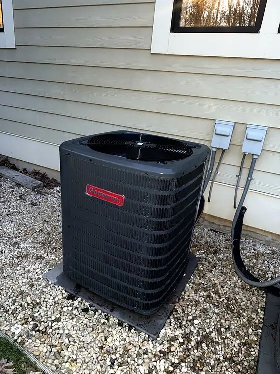 Outdoor air conditioning condenser on a pad in a gravel bed beside tan siding, with two electrical disconnect boxes on the wall.