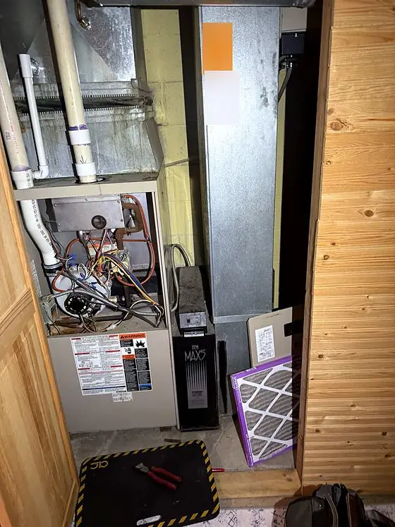 Equipment detail: older furnace with its front panel removed, showing wiring and the burner area, beside an air cleaner cabinet, ductwork and a pleated air filter.