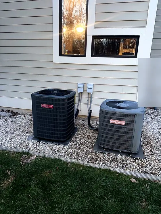 Two outdoor air conditioning condensers side by side on pads in a gravel bed, with electrical disconnects on the siding.
